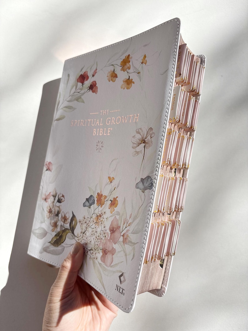 K&ouml;nnte beinhalten: Eine wei&szlig;e Bibel mit floralen Aquarellillustrationen und dem Text "THE SPIRITUAL GROWTH BIBLE". Das Buch hat rosafarbene Seiten und Register. Der Einband zeigt Blumen in Rot, Orange und Blau.