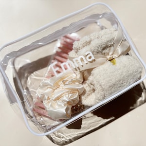 May include: A clear, rectangular cosmetic bag with a white trim, containing various beauty items. Inside are a pink hairbrush, a cream-colored scrunchie, and a fluffy, cream-colored item tied with a ribbon. The name "Emma" is printed on the bag.