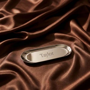 Op de afbeelding: Een gepolijst zilverkleurig ovaal metalen dienblad met de naam Taylor in het midden gegraveerd. Het dienblad heeft een glad, reflecterend oppervlak met opstaande randen en een eenvoudige, langwerpige vorm voor een strakke, metalen uitstraling.