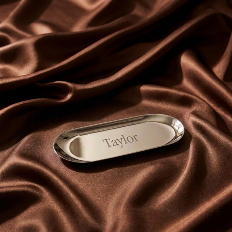 Op de afbeelding: Een gepolijst zilverkleurig ovaal metalen dienblad met de naam Taylor in het midden gegraveerd. Het dienblad heeft een glad, reflecterend oppervlak met opstaande randen en een eenvoudige, langwerpige vorm voor een strakke, metalen uitstraling.