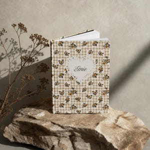 May include: A hardcover book with a beige and white gingham pattern featuring small yellow floral accents. A central white heart-shaped design contains the name Emie in black script. The book is partially open at the top edge.