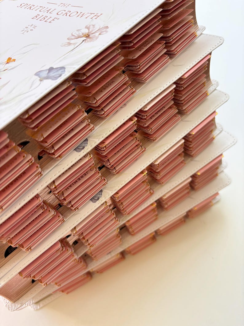 K&ouml;nnte beinhalten: Nahaufnahme einer Bibel mit rosafarbenen Seitenlaschen. Das wei&szlig;e Cover zeigt Blumenillustrationen und den Titel "The Spiritual Growth Bible". Die Laschen sind gestaffelt angeordnet und haben goldene Kanten. Die Seiten sind rosa.