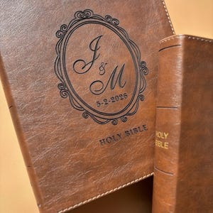 May include: Two brown leather Holy Bibles. The larger Bible has an engraved design with the initials "J & M" and the date "5-2-2026" inside an oval frame. The words "HOLY BIBLE" are printed on the cover.