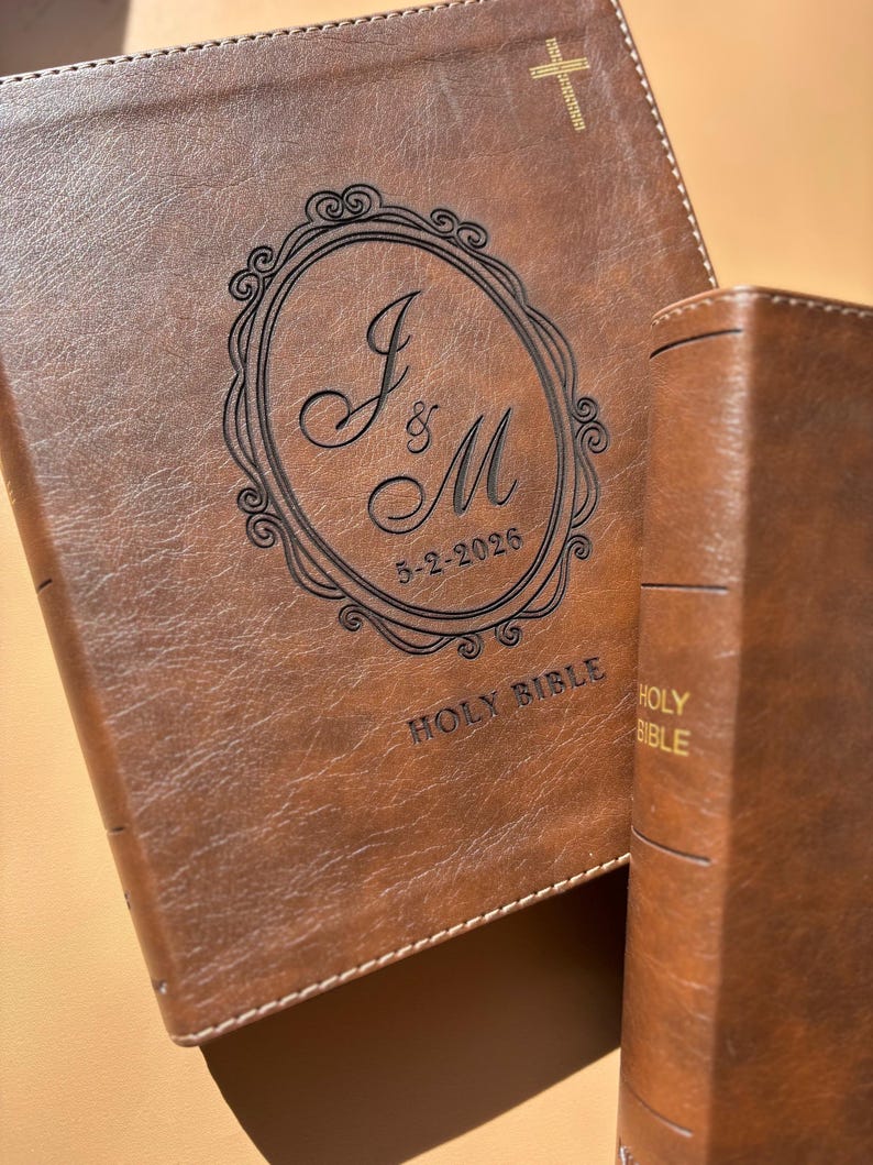 May include: Two brown leather Holy Bibles. The larger Bible has an engraved design with the initials "J & M" and the date "5-2-2026" inside an oval frame. The words "HOLY BIBLE" are printed on the cover.
