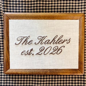 May include: A rectangular wooden frame containing a piece of off-white fabric with brown embroidered text that reads The Kahlers est. 2026. The frame has a smooth, medium-brown finish and surrounds the centered fabric panel in a simple design.