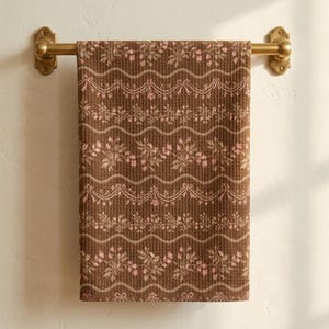 May include: A brown waffle-weave towel hanging on a brass-toned metal bar. The towel features a repeating pattern of horizontal wavy lines, floral sprigs, and decorative ribbon swags in shades of light pink and muted green.