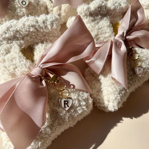May include: Close-up of several fluffy white socks tied with light pink satin ribbons. Each sock has a gold heart detail and a small gold charm with a letter inside a heart. The socks are arranged on a light beige surface.
