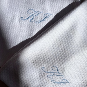 May include: Close-up of white waffle-weave towels with light blue embroidered initials. The towels are folded and arranged, with the initials "KJ" and "JJ" visible. The lighting creates shadows and highlights the texture.