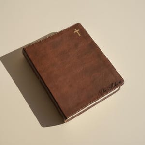 May include: A brown book with a textured cover featuring a small gold cross symbol at the top center and the text The Millers embossed in a cursive script in the bottom right corner. The book has visible white stitching along the edges.