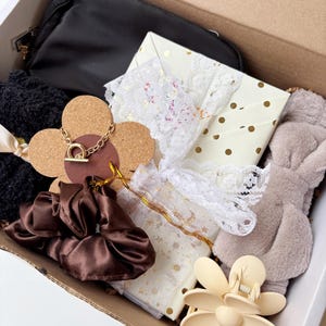 May include: A gift box containing a black pouch, a white box with gold dots and a white lace ribbon, a brown satin scrunchie, a black textured scrunchie, a beige flower-shaped hair claw, a beige plush headband, and a gold necklace on a cork flower.