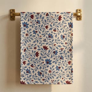 May include: A rectangular waffle-weave kitchen towel featuring a repeating botanical pattern of blue and dark red flowers with green stems and leaves, hanging from a brass-colored metal towel bar mounted on a plain wall.