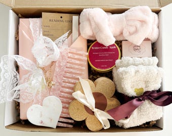 May include: A gift box containing a pink hair comb, a light pink plush headband, a Winter Candy Apple candle, a reading list card, a heart-shaped white stone, a flower-shaped cork coaster with a white ribbon, and fuzzy socks tied with a dark ribbon.
