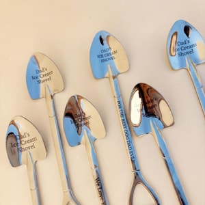 May include: A collection of six silver-toned metal spoons shaped like miniature shovels. Each spoon features engraved text including 'Dad's Ice Cream Shovel', 'FOR THE BEST DOG DAD EVER', 'We Love You', and 'Grandaddy's Ice Cream Spoon' on the bowls and handles.