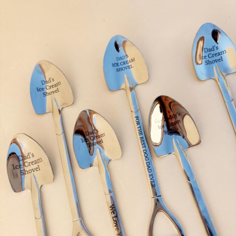 May include: A collection of six silver-toned metal spoons shaped like miniature shovels. Each spoon features engraved text including 'Dad's Ice Cream Shovel', 'FOR THE BEST DOG DAD EVER', 'We Love You', and 'Grandaddy's Ice Cream Spoon' on the bowls and handles.