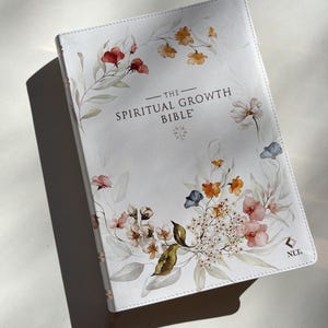 K&ouml;nnte beinhalten: Eine wei&szlig;e Bibel mit floralen Aquarellillustrationen in Rot-, Orange-, Rosa- und Blaut&ouml;nen. Der Titel "The Spiritual Growth Bible" ist in Schwarz gedruckt. Ein hellrosa Lesezeichenband ist sichtbar.
