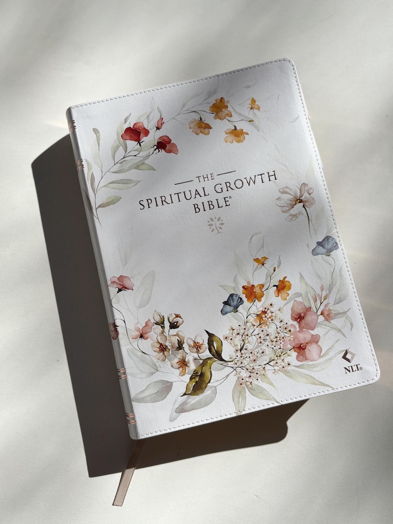 K&ouml;nnte beinhalten: Eine wei&szlig;e Bibel mit floralen Aquarellillustrationen in Rot-, Orange-, Rosa- und Blaut&ouml;nen. Der Titel "The Spiritual Growth Bible" ist in Schwarz gedruckt. Ein hellrosa Lesezeichenband ist sichtbar.