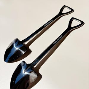 May include: Two silver-colored metal spoons shaped like miniature shovels. Each spoon features a pointed bowl, a long handle, and a triangular grip at the end. The text STAINLESS STEEL is clearly engraved on the handle of each individual spoon.