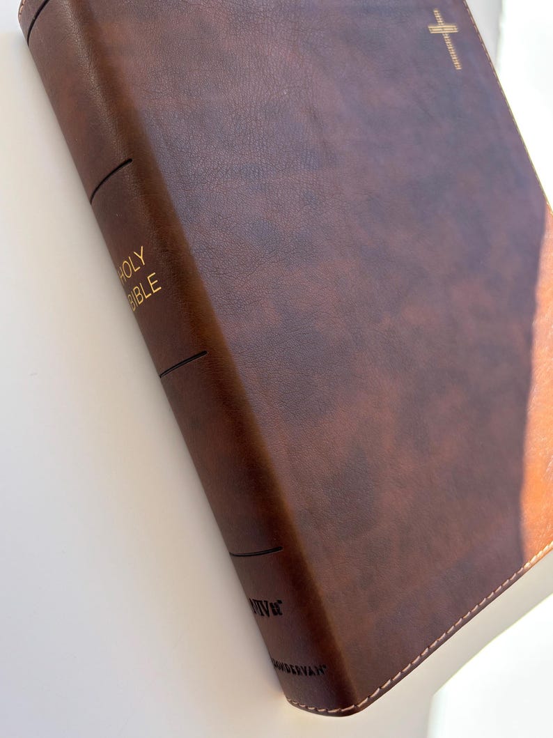 May include: A brown leather-bound Bible with gold lettering that reads "HOLY BIBLE" on the spine. A gold cross is embossed on the cover. The book has a stitched edge.