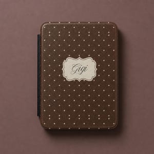 May include: A dark brown e-reader case with a white polka dot pattern and a central cream-coloured decorative label featuring the name Gigi in a black cursive script. The case has a textured finish and a black spine on the left side.