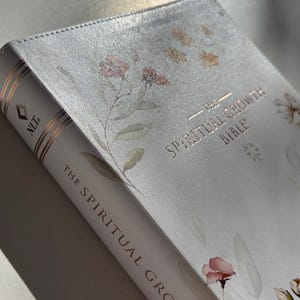 K&ouml;nnte beinhalten: Eine wei&szlig;e Bibel mit floralen Illustrationen und goldfarbenen Buchstaben. Der Titel "The Spiritual Growth Bible" ist auf dem Einband und dem Buchr&uuml;cken zu sehen. Das Buch hat einen weichen, strukturierten Einband mit einem zarten Design.