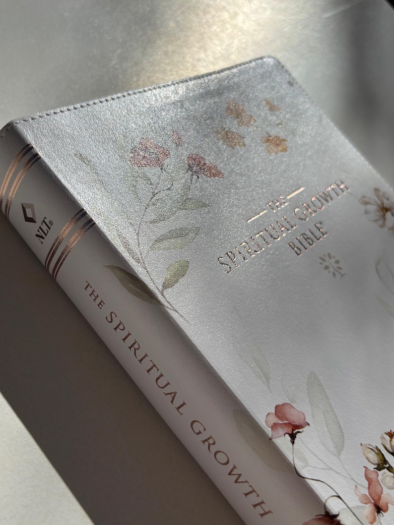 K&ouml;nnte beinhalten: Eine wei&szlig;e Bibel mit floralen Illustrationen und goldfarbenen Buchstaben. Der Titel "The Spiritual Growth Bible" ist auf dem Einband und dem Buchr&uuml;cken zu sehen. Das Buch hat einen weichen, strukturierten Einband mit einem zarten Design.