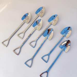 May include: Six silver-toned metal spoons shaped like miniature shovels. Each spoon features a spade-shaped bowl and a D-handle. Engraved text on the bowls includes "Dad's Ice Cream Shovel" and "Daddy's Ice Cream Spoon," with "We Love You Scoopfuls!" on one handle.