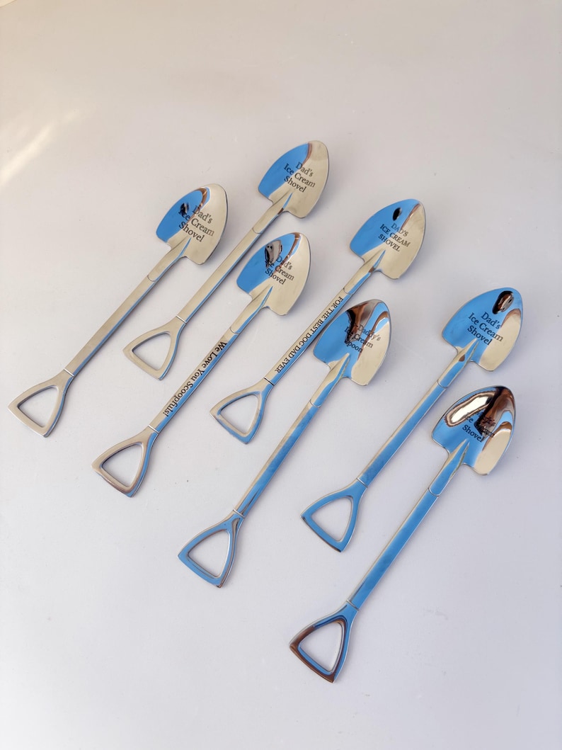May include: Six silver-toned metal spoons shaped like miniature shovels. Each spoon features a spade-shaped bowl and a D-handle. Engraved text on the bowls includes "Dad's Ice Cream Shovel" and "Daddy's Ice Cream Spoon," with "We Love You Scoopfuls!" on one handle.