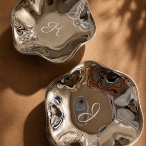 May include: Two silver-toned metal bowls with irregular, wavy edges and a polished, reflective surface. Each bowl features a single white engraved letter in the center, with one displaying the letter K and the other displaying the letter J.