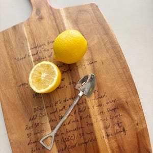 May include: A wooden cutting board with a handle featuring a printed sourdough bread recipe, topped with one whole yellow lemon, one halved yellow lemon, and a silver-colored metal spoon shaped like a miniature shovel with a pointed tip.