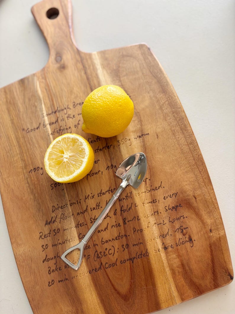 May include: A wooden cutting board with a handle featuring a printed sourdough bread recipe, topped with one whole yellow lemon, one halved yellow lemon, and a silver-colored metal spoon shaped like a miniature shovel with a pointed tip.