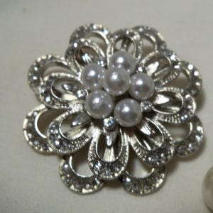 Puede incluir: Broche floral plateado con perlas de imitación en el centro y pedrería transparente alrededor de los pétalos. También se ve un anillo a juego con una perla. Este conjunto de joyas es adecuado para ocasiones especiales.
