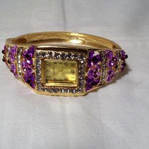 May include: A gold-tone bangle watch featuring a square face with a gold dial and black numerals. The bezel and band are decorated with clear crystals and purple floral-shaped accents. The watch has a hinged design and a quartz movement.
