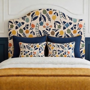 May include: A bed with a floral patterned headboard, navy blue pillows, and decorative throw pillows. The bed is made with a white comforter and a mustard yellow waffle-weave blanket. The room has navy blue wainscoting.