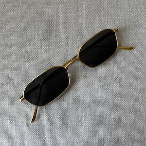Puede incluir: Gafas de sol con una montura fina de metal en tono dorado y lentes rectangulares. Las lentes son oscuras, opacas y cubren completamente el interior de los aros metálicos. Las varillas son delgadas y nacen de las esquinas exteriores.
