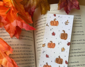 May include: A white bookmark with a repeating pattern of orange pumpkins, leaves, and stars. The bookmark is placed on an open book with fall-colored leaves in the background. The bookmark is protected by a clear plastic sleeve.