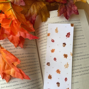 Könnte beinhalten: Ein Lesezeichen mit weißem Hintergrund und Herbstthema. Das Lesezeichen zeigt verschiedene braune und orange Blätter, Eicheln und weiße Punkte. Der Text auf dem Lesezeichen lautet "Es wären nur die Rosen und zerbrochen"