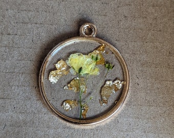 May include: A round pendant with a gold-colored frame. Inside, a yellow flower and gold leaf flakes are encased in clear resin. The pendant has a small loop at the top for a chain. The background is a textured, neutral color.