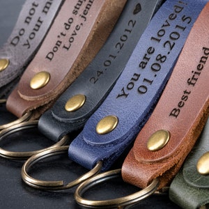 May include: A set of six leather keychains with different engraved messages. The keychains are in various colors, including gray, brown, blue, and green. Each keychain has a brass ring and a brass button.