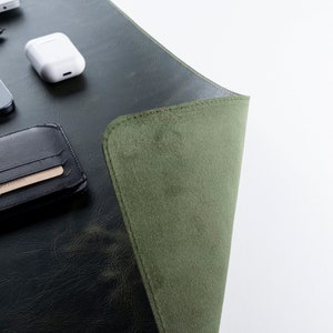 May include: A green leather desk mat with a folded edge, a black leather wallet with a card slot, a black smartphone with a camera lens, and a white wireless charging case.