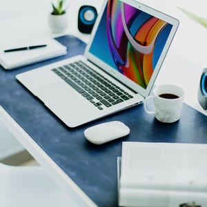 May include: A modern workspace featuring a laptop with a colorful screen, a white mouse, and a marble-patterned coffee mug. The desk has a blue leather desk mat, a notebook, and a pen. A small plant and speakers are also visible.