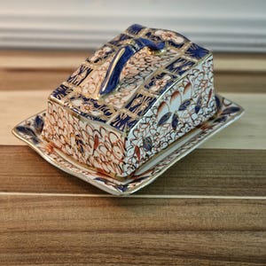 May include: A rectangular ceramic butter dish with a lid, featuring a blue handle and intricate floral patterns in white, orange, and blue. The dish sits on a matching rectangular base with a gold trim, showcasing a vintage aesthetic.