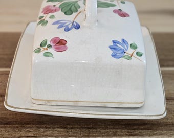 Könnte beinhalten: Eine weiße Keramik-Butterdose mit rechteckigem Deckel und passender Basis. Der Deckel hat einen Griff und ist mit rosa, blauen und grünen Blumendesigns verziert. Die Basis hat einen goldenen Rand.