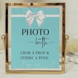 May include: A teal blue and white photo booth sign with a white bow and silver glitter. The sign reads "PHOTO booth" and "GRAB A PROP & STRIKE A POSE".