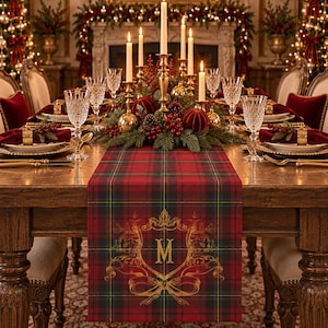 May include: A rectangular table runner featuring a red, green, and black plaid pattern with a gold-coloured ornate crest design in the centre. The crest includes a prominent letter M surrounded by decorative scrollwork and a ribbon bow motif.