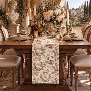 May include: A rectangular table runner featuring a brown and white toile de Jouy pattern with a central crest containing the letter M. The runner is placed on a wooden dining table set with crystal glasses, white plates, and tall candles.