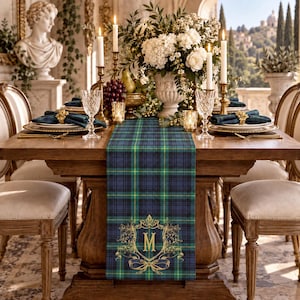 May include: A rectangular table runner featuring a dark blue, green, and yellow plaid pattern. The runner is decorated with a gold-colored ornate crest design centered at the bottom, which features a large letter M surrounded by intricate scrollwork.