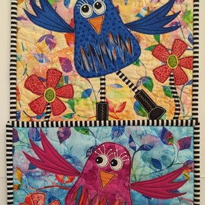 May include: Two colorful quilted wall hangings featuring whimsical birds with large eyes and boots. The top bird is blue with orange beak and the bottom bird is pink with orange beak. Both birds have black and white striped legs and are surrounded by flowers and green grass.