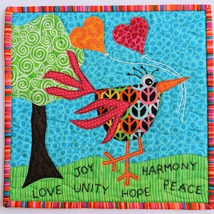 May include: A colorful quilted wall hanging featuring a whimsical bird with peace signs on its body, holding balloons in the shape of hearts. The background is a bright blue with a green tree and the words "LOVE UNITY JOY HOPE HARMONY PEACE" are stitched on the bottom.