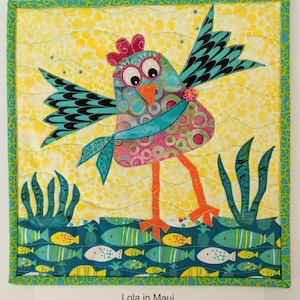 Könnte beinhalten: Ein farbenfroher, gesteppter Wandbehang mit einem Cartoon-Vogel mit blauem Schal, der auf einem blauen und gelben Hintergrund mit Fischen steht. Der Text "Lola in Maui" steht unter dem Bild.