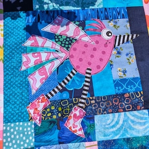 May include: A pink and white bird applique with black and white striped legs, surrounded by blue and green fabric squares. The bird has a large, round eye and a black and white striped beak.